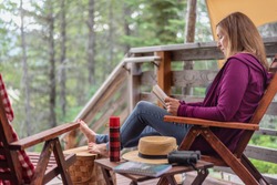 glamping trip - woman relaxing outside cabin in the woods with a book on vacation