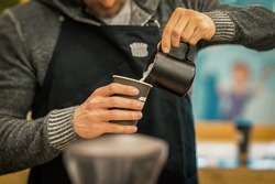 Unrecognizable Barista wearing black apron making cappuccino close-up, pouring milk in paper cup in coffee bar