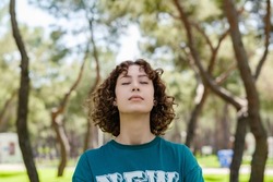 Young redhead woman wearing green tee standing on green city park, outdoors. Enjoying stress free mindful moment, doing yoga relaxation exercises, closed eyes, meditating near nature, taking breath.