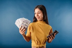 Brown-haired woman wearing mustard yellow t-shirt isolated over blue background holding money banknotes, using mobile phone. Showing bunch of money banknotes.