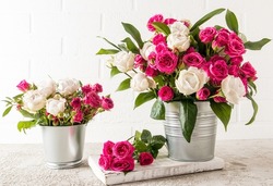 two fresh garden bouquets of roses in decorative buckets stand on a white wooden podium. grey concrete table. white brick wall-background