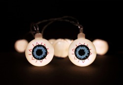 Halloween garland lights of glowing eyeballs on black background with copy space. Blurred white bokeh lights, two eyes in focuse, close up. Festive holidays Halloween decor concept.