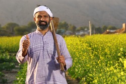 Beautiful dramatic portrait of Indian rural happy farmer standing in mustard field wearing kurta pajama in summer time. Pleased farmer is holding shovel in hand. Flourishing crops.
