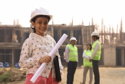 Aspirational happy Indian girl child standing on a construction site holding blue prints and file and wearing engineer's helmet. Portraying to be a future engineer, all the engineers standing behind.