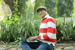 Happy Indian old man sitting in a lush green atmosphere in a park holding a laptop and working online, making the best use of technology and internet. Wearing spectacles and a golf cap.
