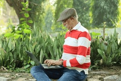 Happy Indian old man sitting in a lush green atmosphere in a park holding a laptop and working online, making the best use of technology and internet. Wearing spectacles and a golf cap.