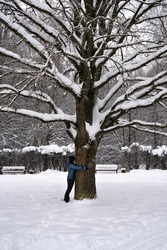 Woman hugs a trunk of a large old oak in a snowy winter park in cold weather
