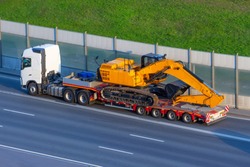 Heavy new yellow excavator on transportation truck with long trailer platform on the highway in the city