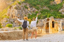 Guy with backpack in hat girl, travelers raising their hands up in surprise, look at ruins ancient city, ancient unique famous places of Turkey, Antalya Demre, Mira, ancient Lycian tomb casts faces