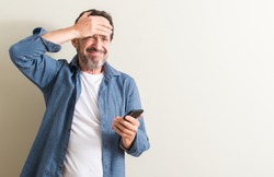 Senior man using smartphone stressed with hand on head, shocked with shame and surprise face, angry and frustrated. Fear and upset for mistake.
