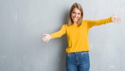 Beautiful young woman standing over grunge grey wall looking at the camera smiling with open arms for hug. Cheerful expression embracing happiness.