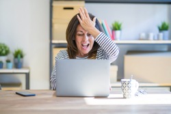 Middle age senior woman sitting at the table at home working using computer laptop surprised with hand on head for mistake, remember error. Forgot, bad memory concept.