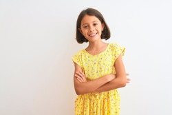 Young beautiful child girl wearing yellow floral dress standing over isolated white background happy face smiling with crossed arms looking at the camera. Positive person.