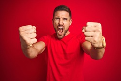 Young handsome man wearing casual t-shirt over red isolated background angry and mad raising fists frustrated and furious while shouting with anger. Rage and aggressive concept.