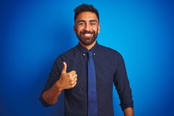 Young indian businessman wearing elegant shirt and tie standing over isolated blue background doing happy thumbs up gesture with hand. Approving expression looking at the camera showing success.