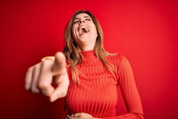 Young beautiful brunette woman wearing casual turtleneck sweater over red background laughing at you, pointing finger to the camera with hand over body, shame expression
