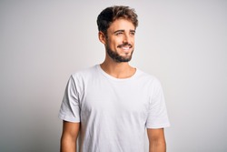 Young handsome man with beard wearing casual t-shirt standing over white background looking away to side with smile on face, natural expression. Laughing confident.