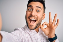 Young handsome man wearing shirt making selfie by the camera over white background doing ok sign with fingers, excellent symbol