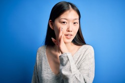 Young beautiful asian woman wearing casual sweater standing over blue isolated background hand on mouth telling secret rumor, whispering malicious talk conversation