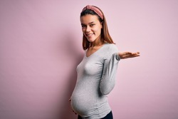 Young beautiful teenager girl pregnant expecting baby over isolated pink background smiling cheerful presenting and pointing with palm of hand looking at the camera.