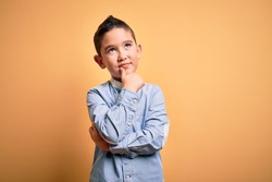 Young little boy kid wearing elegant shirt standing over yellow isolated background with hand on chin thinking about question, pensive expression. Smiling with thoughtful face. Doubt concept.