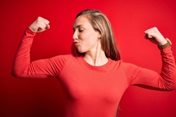 Young beautiful blonde woman wearing casual t-shirt standing over isolated red background showing arms muscles smiling proud. Fitness concept.