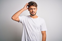 Young handsome man with beard wearing casual t-shirt standing over white background confuse and wonder about question. Uncertain with doubt, thinking with hand on head. Pensive concept.