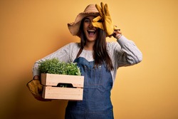 Young beautiful brunette gardener woman wearing apron and hat holding box with plants with happy face smiling doing ok sign with hand on eye looking through fingers