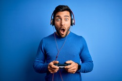 Young handsome gamer man with beard playing video game using joystick and headphones scared in shock with a surprise face, afraid and excited with fear expression