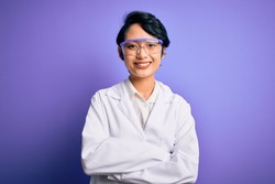 Young beautiful asian scientist girl wearing coat and glasses over purple background happy face smiling with crossed arms looking at the camera. Positive person.