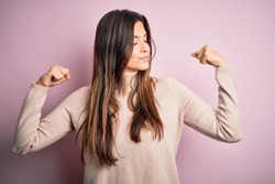 Young beautiful girl wearing casual turtleneck sweater standing over isolated pink background showing arms muscles smiling proud. Fitness concept.