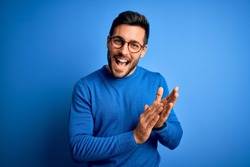 Young handsome man with beard wearing casual sweater and glasses over blue background clapping and applauding happy and joyful, smiling proud hands together