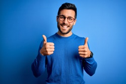 Young handsome man with beard wearing casual sweater and glasses over blue background success sign doing positive gesture with hand, thumbs up smiling and happy. Cheerful expression and winner 