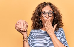 Middle age brunette woman holding brain as mental care and memory health over pink background covering mouth with hand, shocked and afraid for mistake. Surprised expression