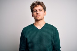 Young blond handsome man with curly hair wearing green sweater over white background Relaxed with serious expression on face. Simple and natural looking at the camera.