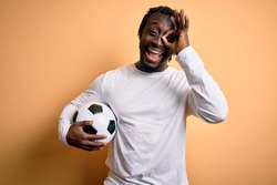 Young african american player man playing soccer holding football ball over yellow background with happy face smiling doing ok sign with hand on eye looking through fingers