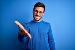Young handsome man with beard wearing casual sweater and glasses over blue background smiling cheerful offering palm hand giving assistance and acceptance.