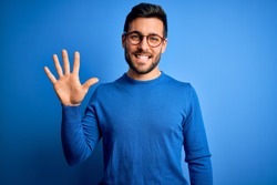 Young handsome man with beard wearing casual sweater and glasses over blue background showing and pointing up with fingers number five while smiling confident and happy.