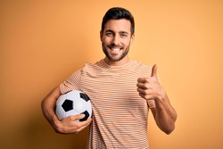 Handsome player man with beard playing soccer holding footballl ball over yellow background doing happy thumbs up gesture with hand. Approving expression looking at the camera showing success.