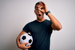 Handsome african american man playing footbal holding soccer ball over white background doing ok gesture shocked with surprised face, eye looking through fingers. Unbelieving expression.