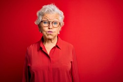Senior beautiful grey-haired woman wearing casual shirt and glasses over red background puffing cheeks with funny face. Mouth inflated with air, crazy expression.