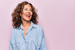 Middle age beautiful woman wearing casual denim shirt standing over pink background looking away to side with smile on face, natural expression. Laughing confident.
