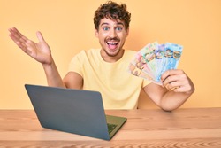 Young caucasian man with curly hair sitting on the table working with laptop and holding canadian dollars banknotes celebrating victory with happy smile and winner expression with raised hands 