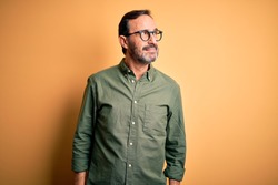 Middle age hoary man wearing casual green shirt and glasses over isolated yellow background looking away to side with smile on face, natural expression. Laughing confident.