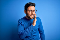 Young handsome man with beard wearing casual sweater and glasses over blue background hand on mouth telling secret rumor, whispering malicious talk conversation