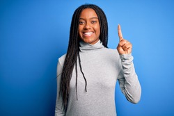 Young african american woman standing wearing casual turtleneck over blue isolated background showing and pointing up with finger number one while smiling confident and happy.