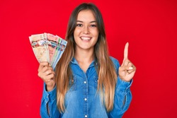 Beautiful caucasian woman holding canadian dollars smiling with an idea or question pointing finger with happy face, number one 