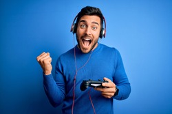 Young handsome gamer man with beard playing video game using joystick and headphones screaming proud and celebrating victory and success very excited, cheering emotion