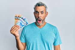 Middle age grey-haired man holding canadian dollars scared and amazed with open mouth for surprise, disbelief face 