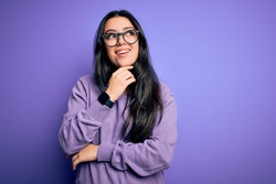 Young brunette woman wearing glasses over purple isolated background with hand on chin thinking about question, pensive expression. Smiling with thoughtful face. Doubt concept.